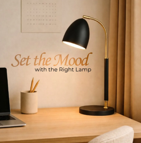 A modern desk lamp with a black dome-shaped shade and a curved gold metal stem on a round black base, placed on a wooden desk in a styled home office setting. The scene includes a laptop, a pencil cup, a calendar on the wall, and the text 'Set the Mood with the Right Lamp'.